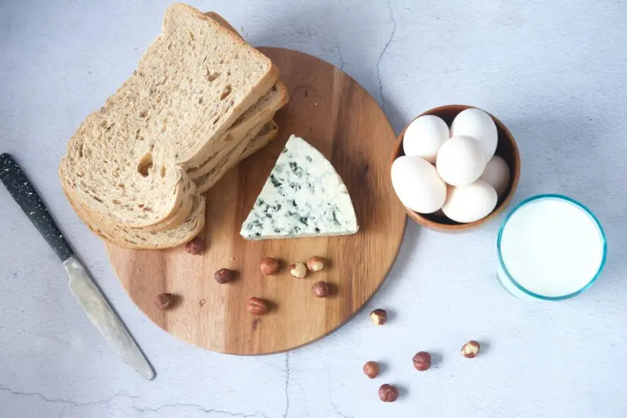 Image of a wooden board with a block of cheese next to slices of bread, eggs, and a glass of milk.