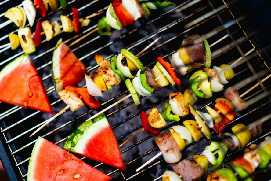 Image of several fruit and vegetable skewers shown on a barbecue grill alongside slices of watermelon.