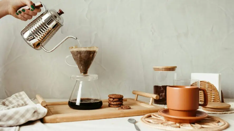 Image of a hand pouring water from a metal kettle into a coffee pour-over set-up, resting on a wooden plank with brown cookies alongside it.