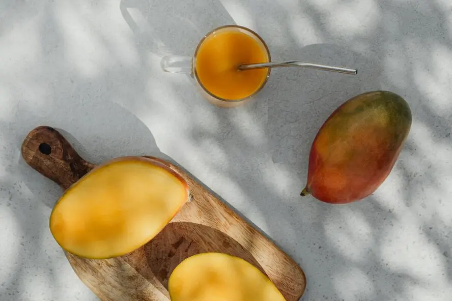 Image of a couple of mangoes (one whole, one sliced) shown on a cutting board and white tablecloth next to a smoothie.