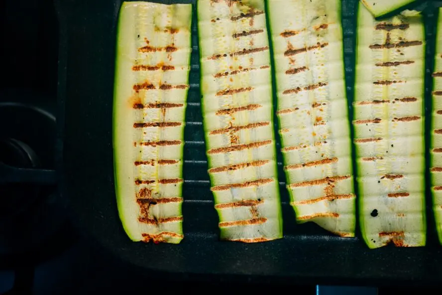 Image of four thin strips of zucchini shown on a grill, lightly charred with grill marks.