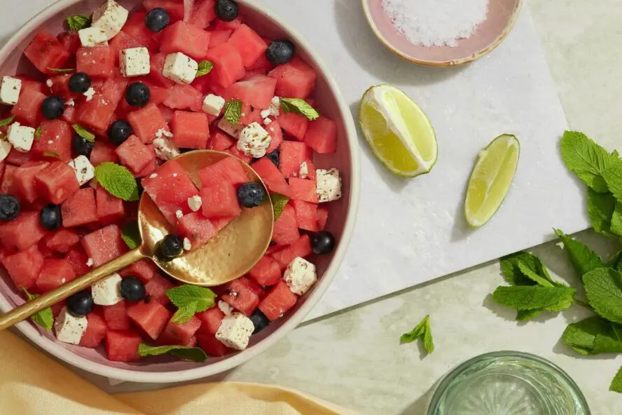 Image of a large bowl of watermelon and feta salad, shown on a cutting board with wedges of lime.