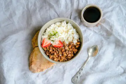 Image of a bowl of cottage cheese topped with granola and strawberries, served on a rustic wooden plank next to a spoon and a cup of black coffee.
