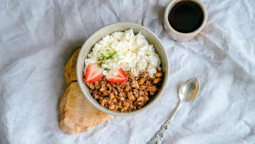 Image of a bowl of cottage cheese topped with granola and strawberries, served on a rustic wooden plank next to a spoon and a cup of black coffee.