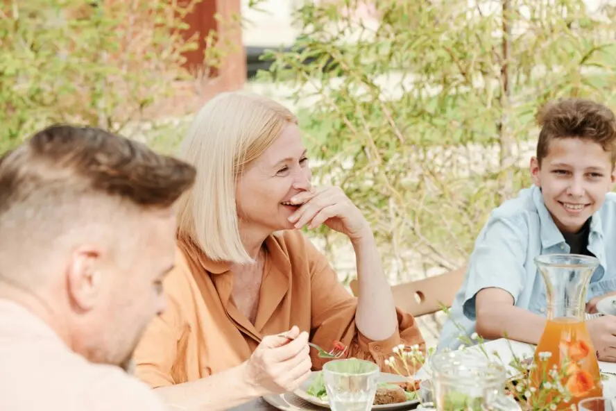Image of an older woman sitting outside at a table with her family, enjoying a meal together.