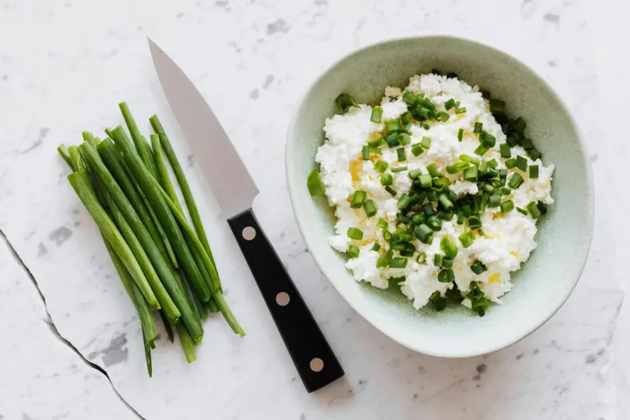Image of a small bowl of cottage cheese topped with tiny slivers of chives, shown on a marble countertop next to a knife.