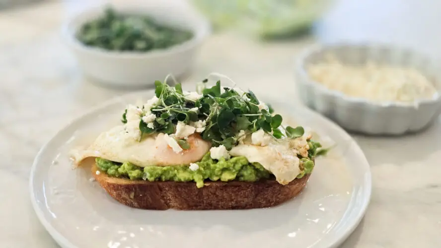 Image of a thick slice of toasted sourdough topped with mashed avocado, butter-basted eggs, microgreens, and feta.