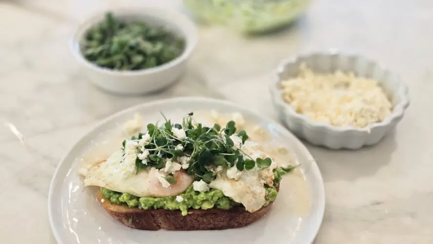 Image of a slice of toast topped with avocado and eggs, with small dishes of microgreens and feta behind the plate.