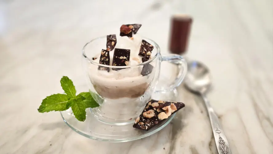 Image of vanilla bean gelato in a glass cup on a small dish, topped with chocolate hazelnut bark alongside a sprig of mint, a spoon, and a shot of espresso.