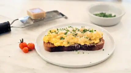 Image of a thick slice of sourdough topped with soft scrambled eggs, Parmesan cheese, and thinly sliced chives shown on a marble countertop.