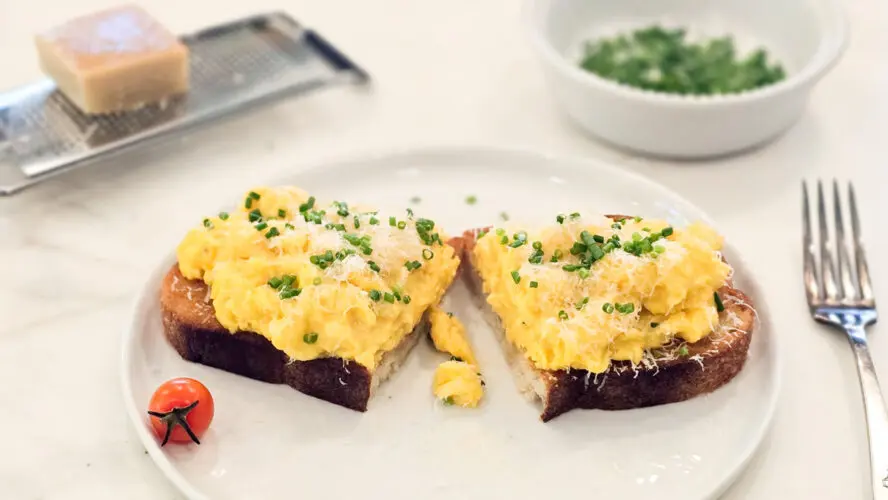 Image of a piece of toast topped with French scrambled eggs, sliced in half on a plate.