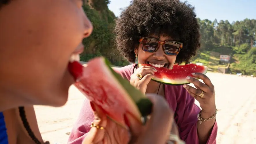 Image of two young women at the beach smiling as they eat large slices of watermelon.