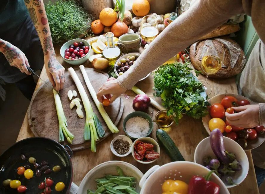 Image of a brightly colored spread of food, mostly plant-based, shown on a wooden table with hands reaching over it.
