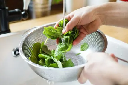 Image of a pair of hands washing spinach under running water in a sink with a colander below it.