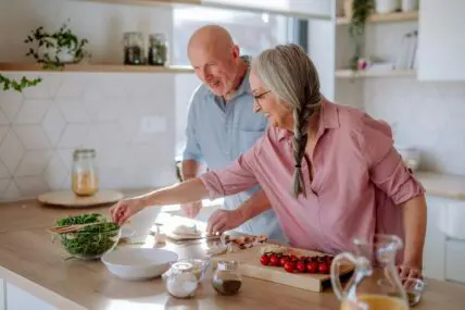 Image of a senior couple in a brightly lit kitchen, standing at the counter and beginning to prepare a meal together.