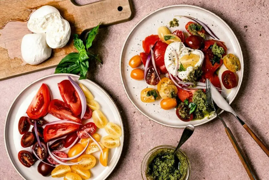 Image of two plates full of vibrant tomatoes, onion, basil, pesto, and other fresh ingredients.