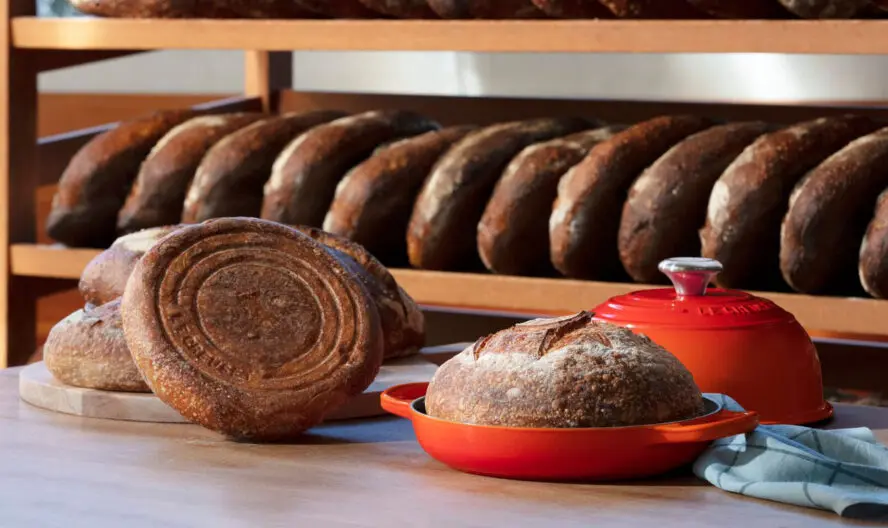 Image of a Le Creuset Cast Iron Bread Oven in the color Volcanic, shown with a number of bread loaves behind it.