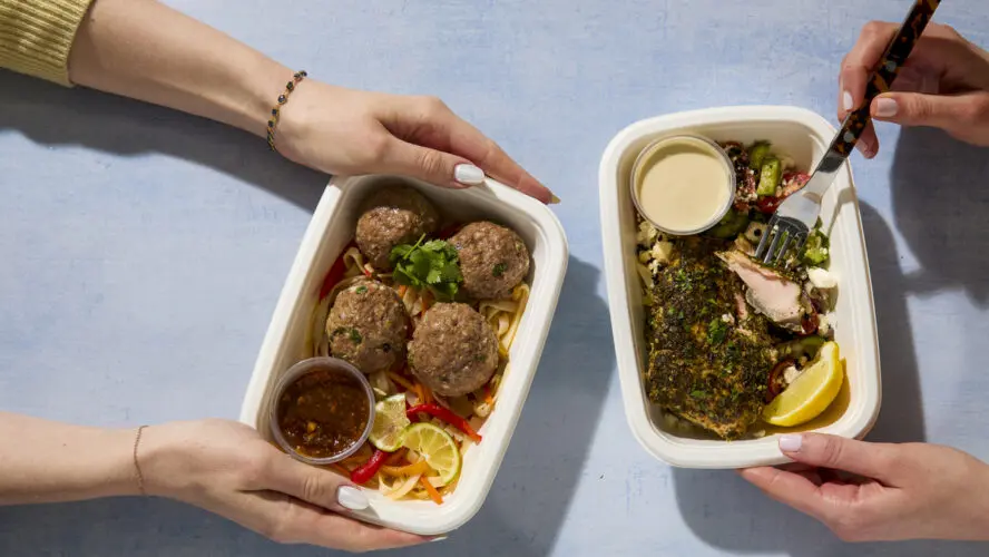 Image of two pairs of hands holding CookUnity meal trays with different meals shown.
