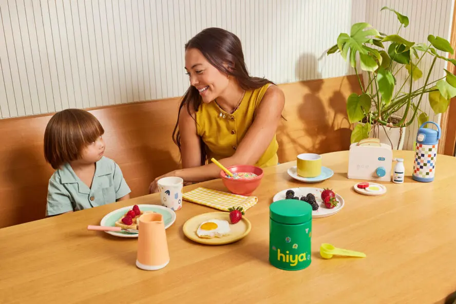 Image of a young mom and child sitting at a wooden table, with breakfast and a bottle of Hiya Kids Vitamins in front of them.