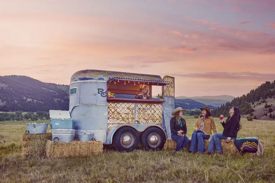 Image of a cute outdoor bar run out of a trailer, with three women sitting in front of it enjoying drinks at sunset.
