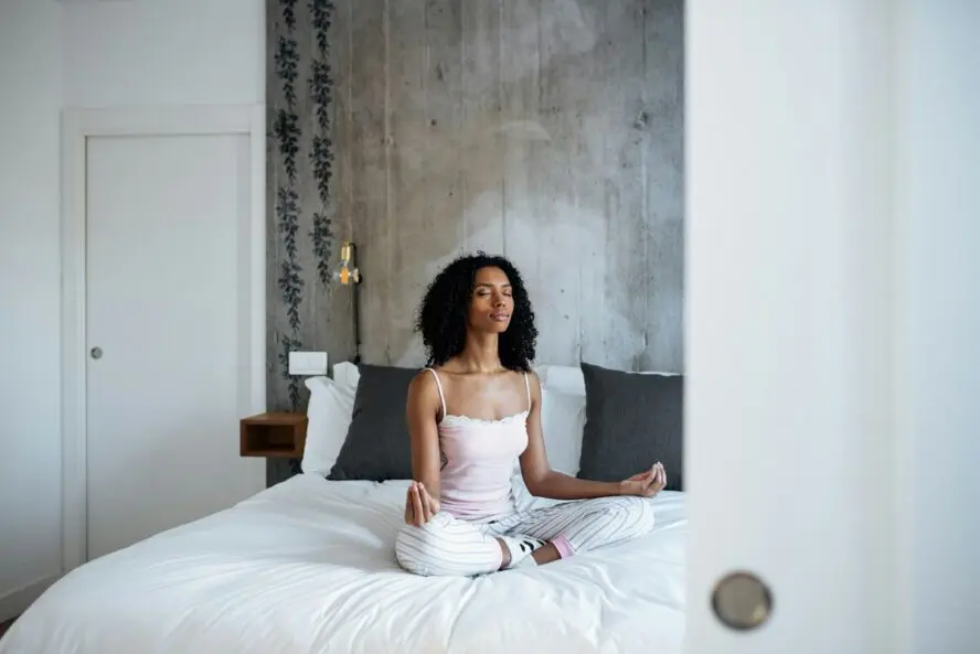 Image of a young woman sitting cross-legged on a bed with her hands resting on her knees and her eyes closed in meditation.