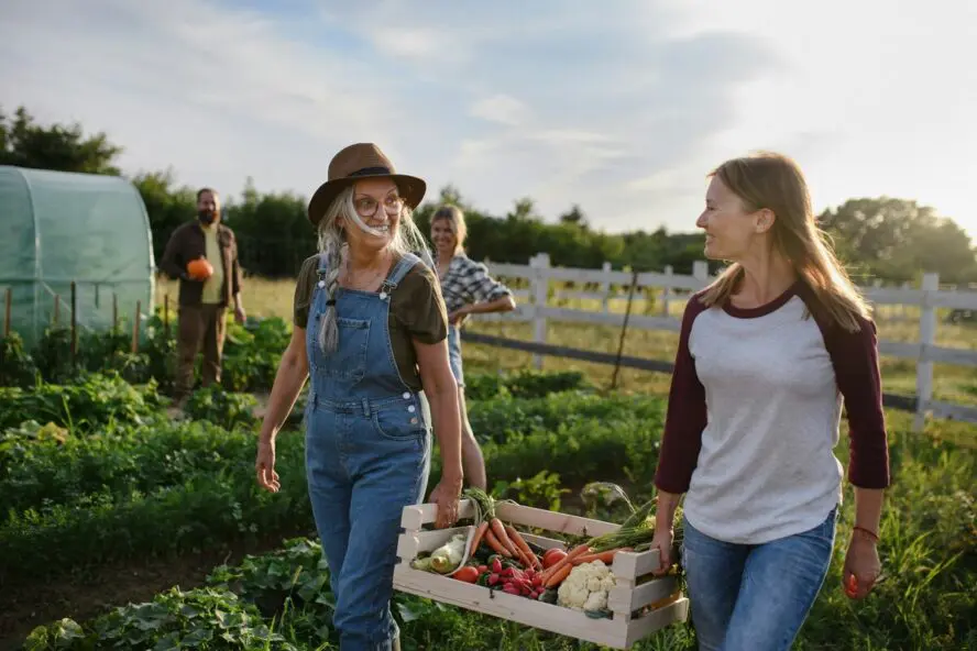 Image of a younger woman and an older woman on a farm, holding a pallet full of fresh produce.