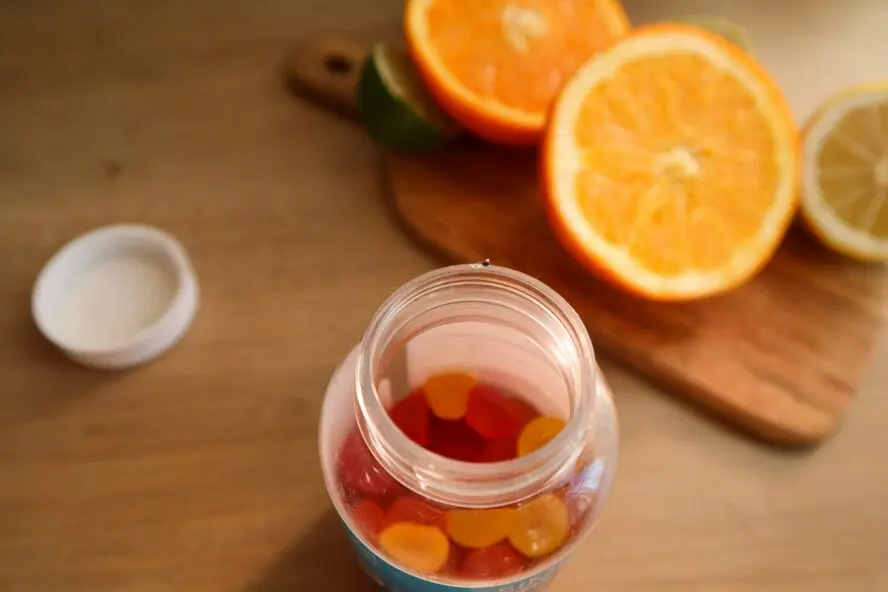 Image of a plastic bottle of gummy vitamins, shown with the lid off next to an orange sliced in half.