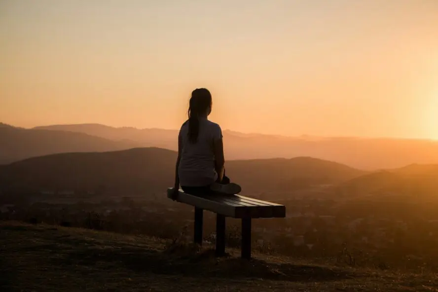 Image of a woman sitting on a bench and looking out into the distance at sunset.