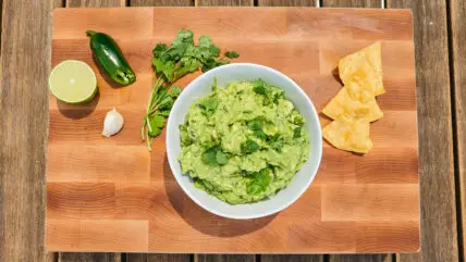 bowl of fresh homemade guacamole topped with cilantro on a wood cutting board with a halved lime and jalapeño and three tortilla chips.