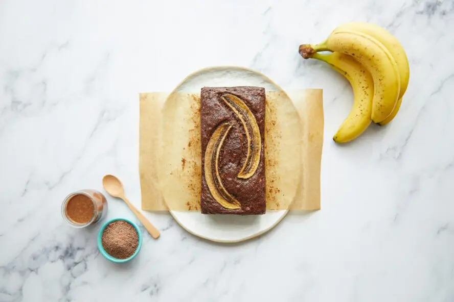 Image of a plate with parchment paper and a loaf of banana bread, set on a marble table next to two bananas and other baking ingredients.
