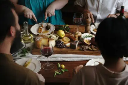 Image of a group of people sitting at a dinner table covered in various cheeses and glasses of red wine.