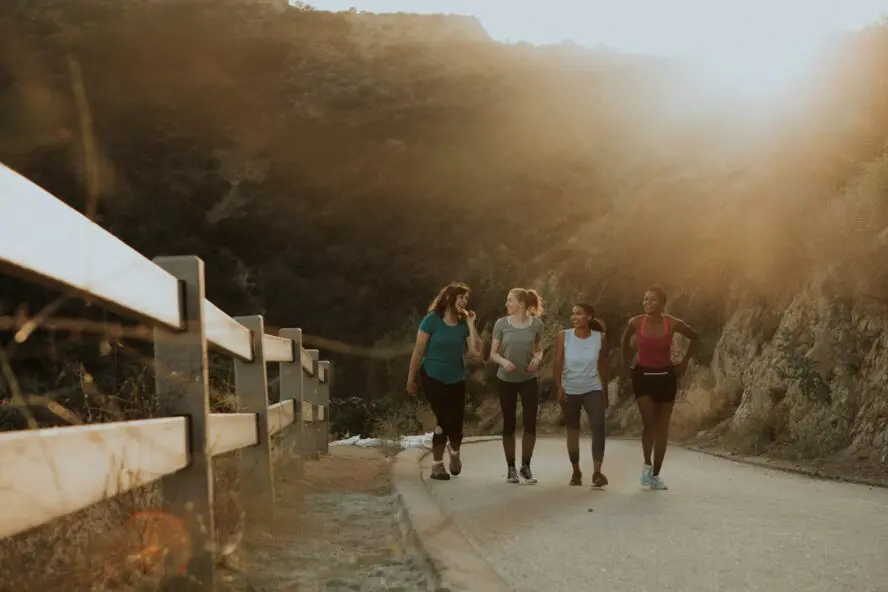 four women walking on trail next to a fence with glowing sun setting in background