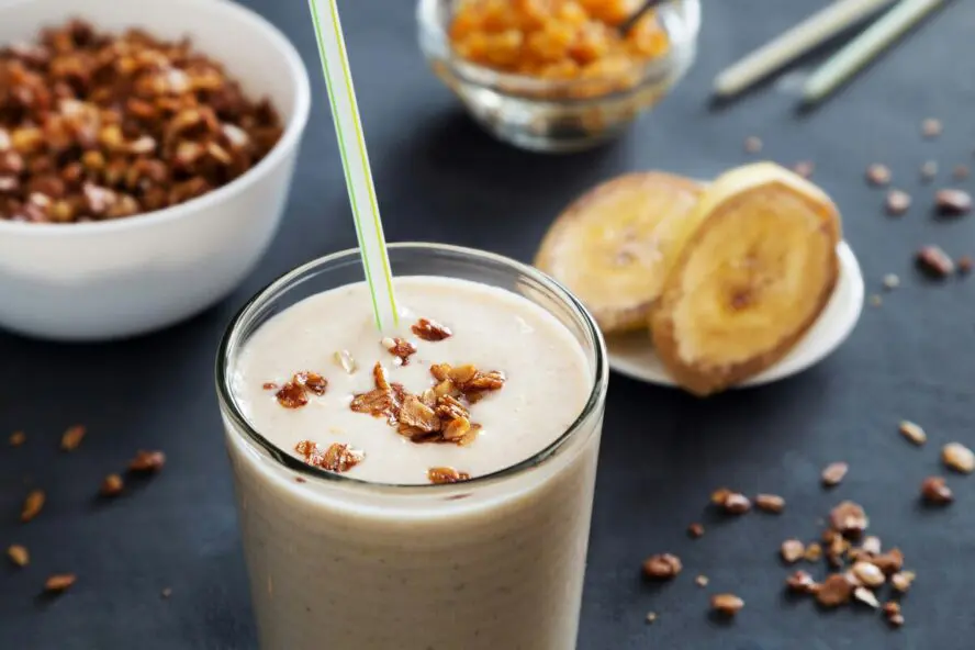 Image of a banana smoothie shown next to slices of banana and a crunchy granola topping.