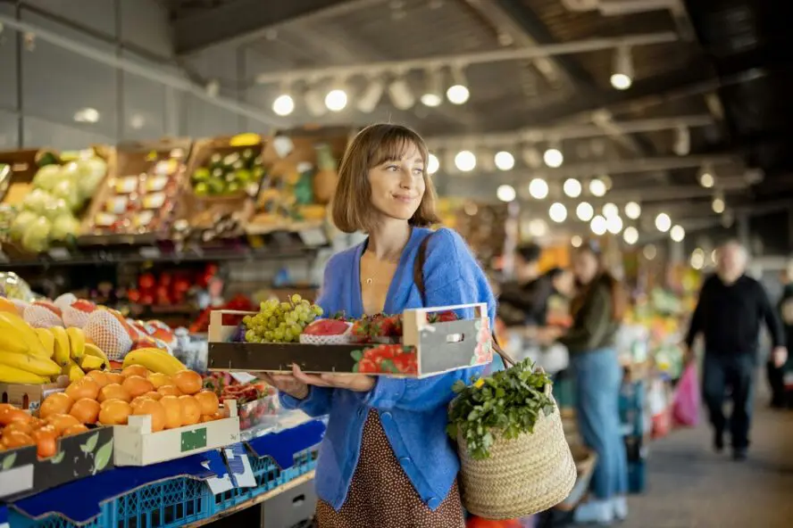 Image of a young woman in a blue cardigan holding a box full of fresh fruit and vegetables in her arms at the grocery store.