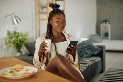 black woman holding phone at home while having breakfast and holding glass of milk.
