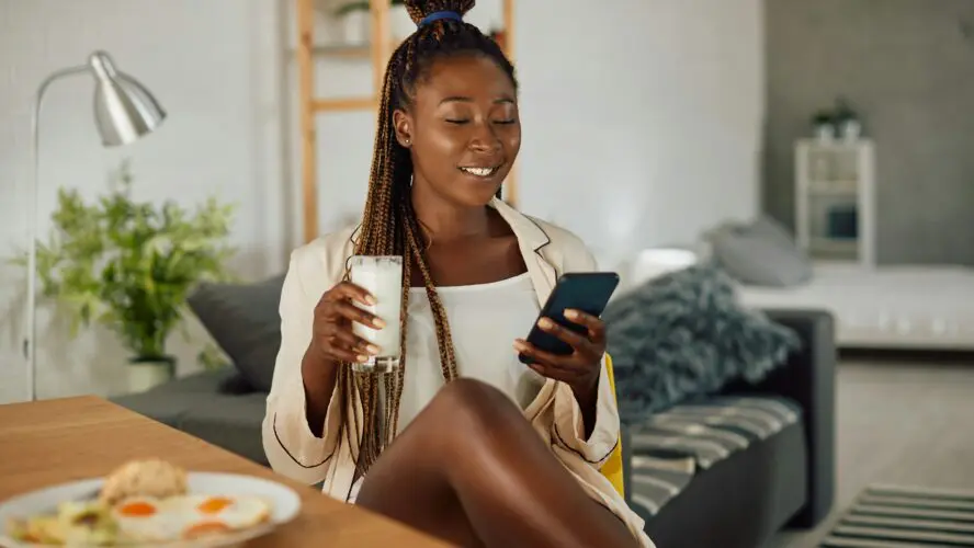 black woman holding phone at home while having breakfast and holding glass of milk.