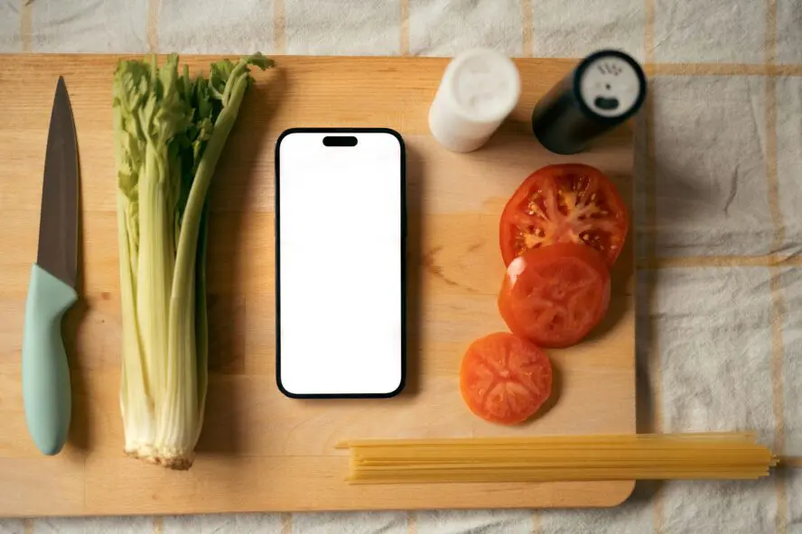 lit up phone screen on cutting board with fresh sliced tomato, a celery heart, and salt and pepper