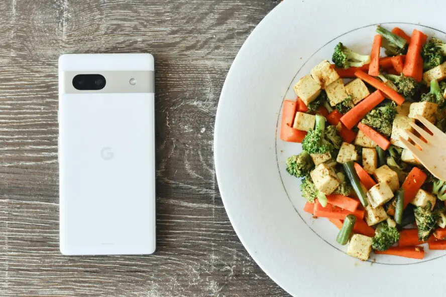 white plate of tofu stir fry with vegetables alongside an Android phone face down on wood surface