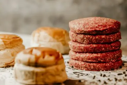 Image of several raw ground beef burgers stacked on top of each other next to buns.