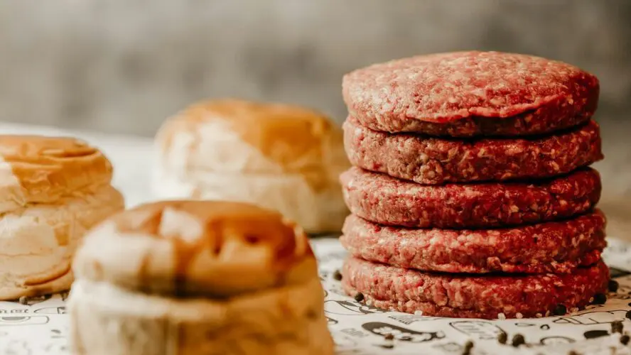 Image of several raw ground beef burgers stacked on top of each other next to buns.