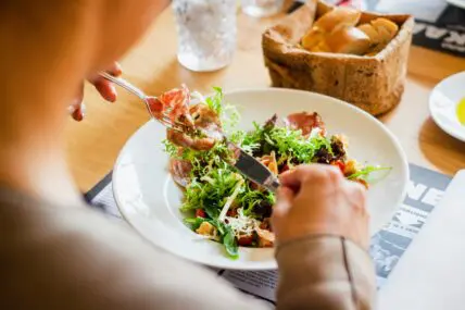 Image of a a bowl of salad with person holding fork with some food on it and a knife.