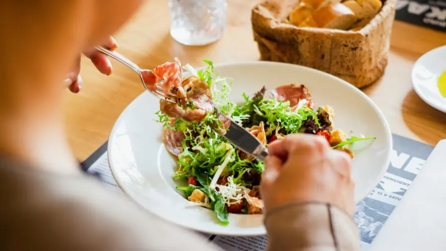 Image of a a bowl of salad with person holding fork with some food on it and a knife.