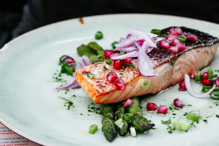 salmon with crispy skin on a white plate over a bed of greens, topped with sliced red onions and pomegranate arils. 