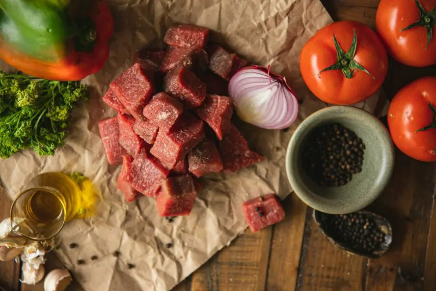 Image of a tabletop covered with parchment paper and thick chunks of raw beef, alongside pepper, an onion, and tomatoes.