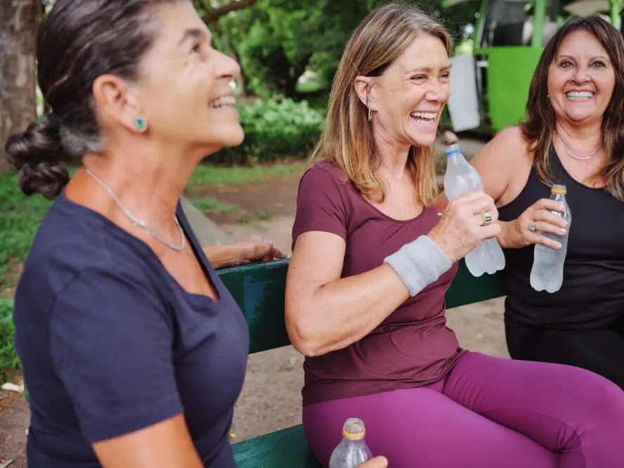 Image of three middle-aged women sitting on a bench and drinking water after a workout.
