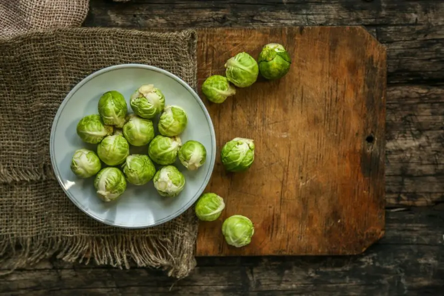 Image of a small plate full of raw Brussels sprouts, with a wooden cutting board next to it waiting to be prepped.