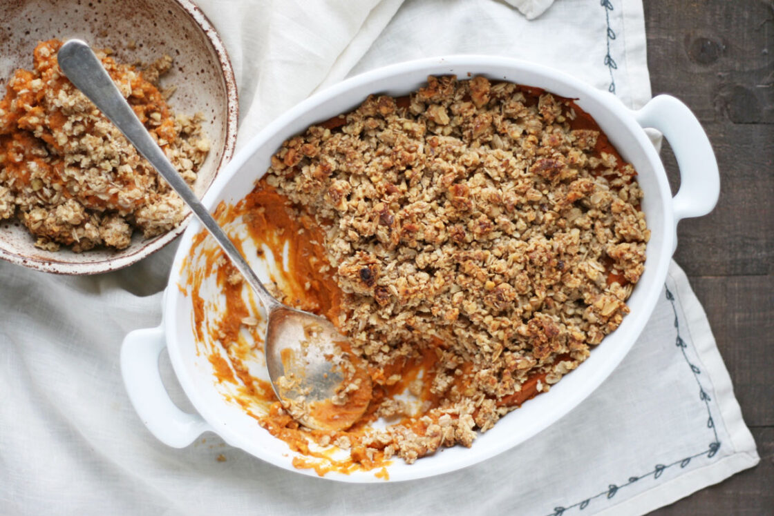 Image of sweet potato casserole topped with an oat crumble, served in white ceramic bakeware on white towel alongside a bowl of a serving