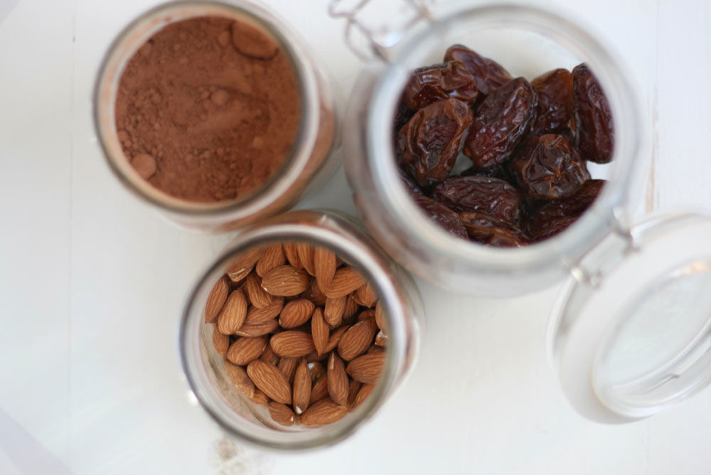 Image of 3 glass canisters of ingredients for no bake peanut butter cookies with chocolate, cocoa powder, dates, and almonds on a white countertop.