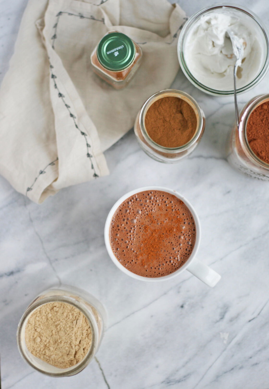 Image of overhead view of Vegan Superfood cacao Hot Chocolate With Reishi, Maca, and Raw Cacao on white marble countertop with ingredients and coconut whipped crea.