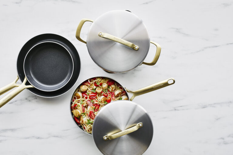 Image of an overhead view of a set of the best ceramic cookware by GreenPan in their GP5 line, with a saucepan filled with pasta with its lid askew showing what's inside. It's ceramic-coated nonstick with a beautiful stainless steel finish and champagne handles.